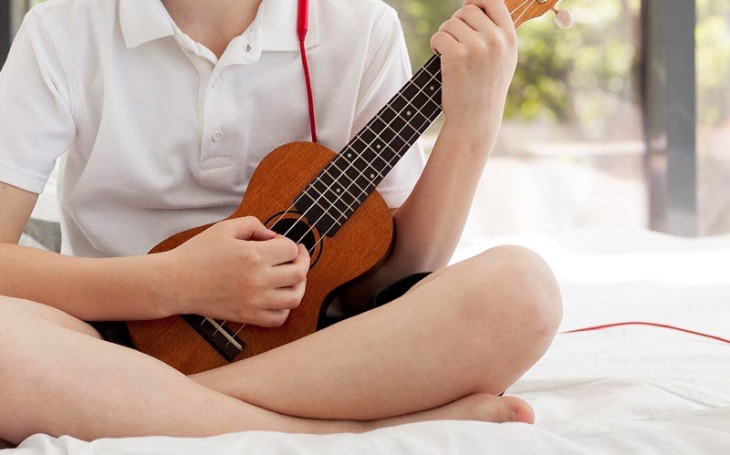 Child holding a wooden soprano ukulele on a bed.