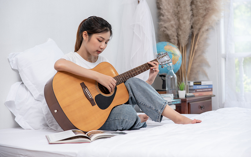 Teenager practicing acoustic guitar on a bed.